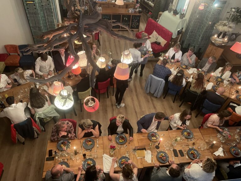 Gezelschap geniet van diner in een sfeervol restaurant met hangende lampen aan een houten constructie.