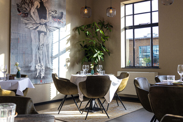 Zonnig restaurant met gedekte ronde tafels, olijfgroene fluwelen stoelen, grote plant en ballerina schilderij.