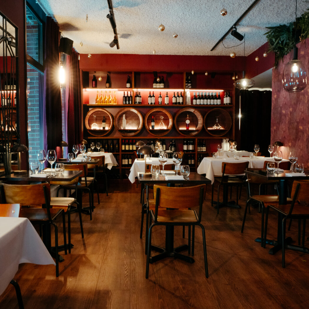 Gezellig restaurant interieur met gedekte tafels, houten vloer en een rode muur met verlichte wijnrekken en vaten.