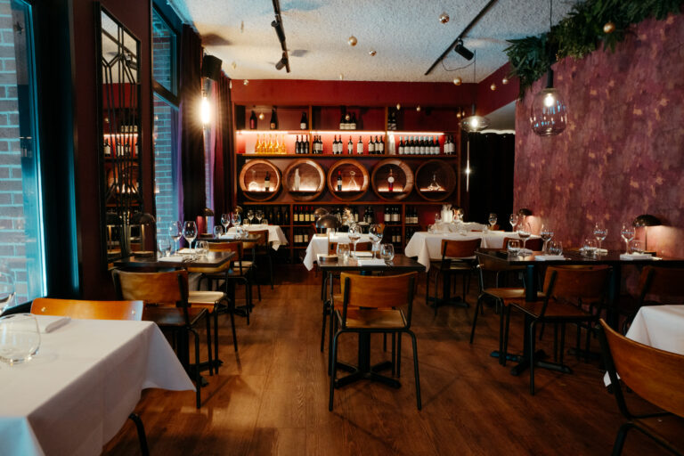 Gezellig restaurant interieur met gedekte tafels, houten vloer en een rode muur met verlichte wijnrekken en vaten.
