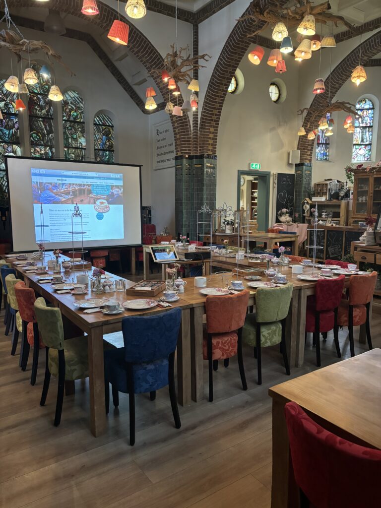 Ruime evenementenlocatie met U-vormige houten tafel, kleurrijke stoelen, projector, glas-in-lood en stoffen hanglampen.