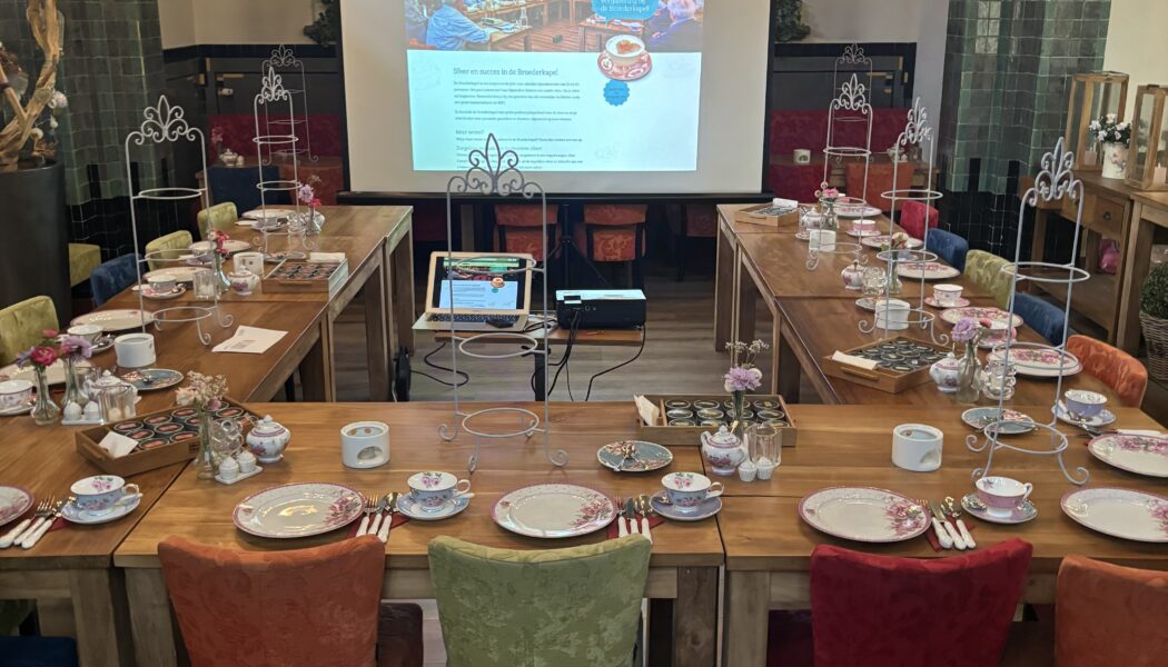 Ruimte in een kapel met glas-in-loodramen en U-vormige tafels, gedekt voor high tea, met een projectie van Meer en succes in de Brinkkapel.
