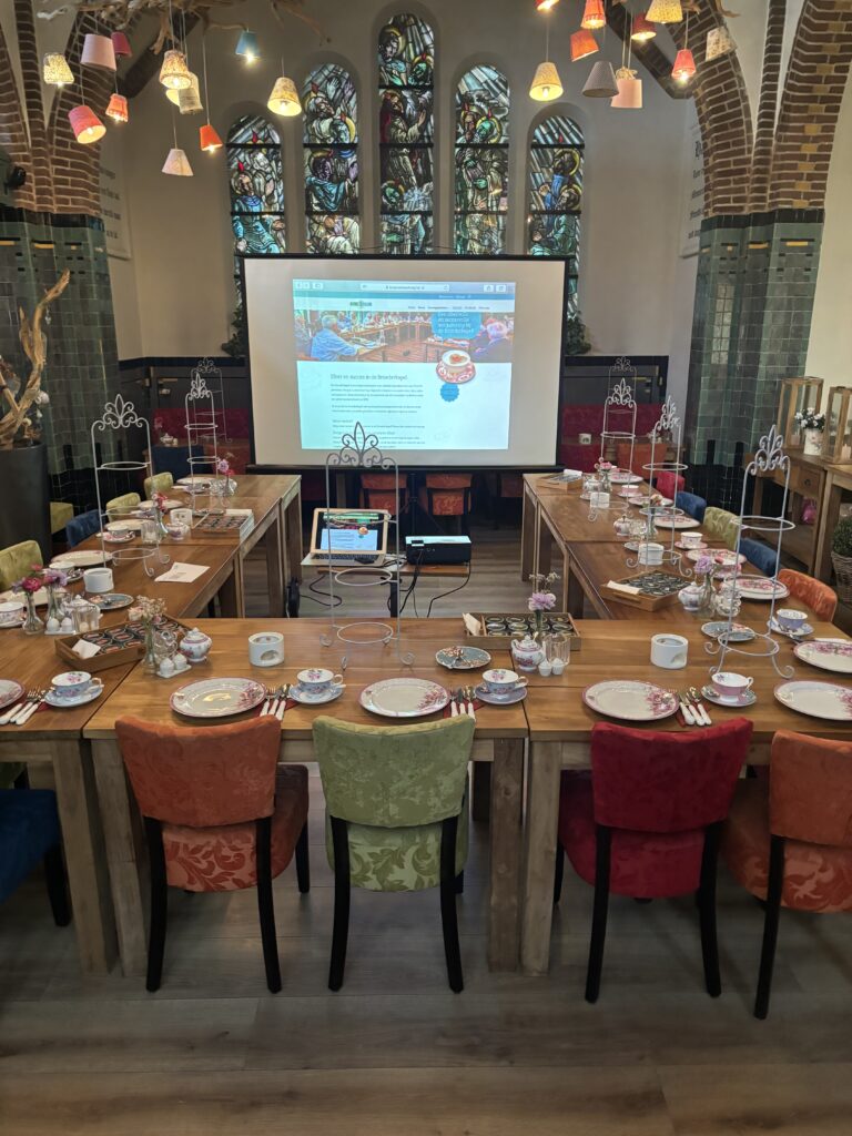 Ruimte in een kapel met glas-in-loodramen en U-vormige tafels, gedekt voor high tea, met een projectie van Meer en succes in de Brinkkapel.