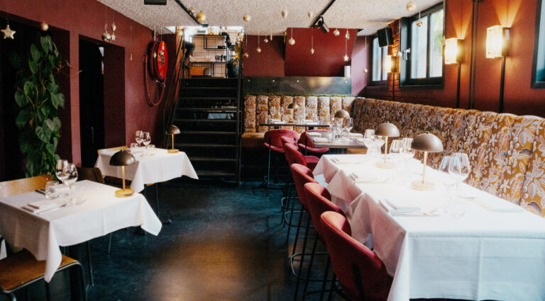 Sfeervol restaurantinterieur met rode muren, wit gedekte tafels, fluwelen stoelen en feestelijke plafonddecoraties.