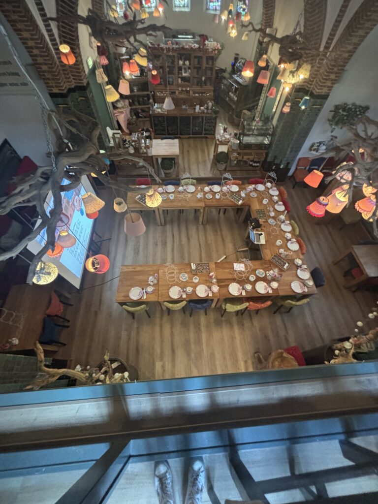 Bovenaanzicht van een sfeervol theesalon met lange U-vormige tafel gedekt voor high tea en kleurrijke lampenkappen.