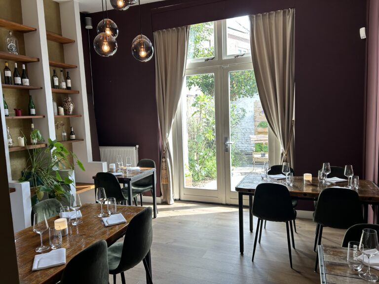 Warm restaurantinterieur met gedekte houten tafels, donkergroene stoelen, sfeervolle verlichting en een grote raampartij naar een tuin.