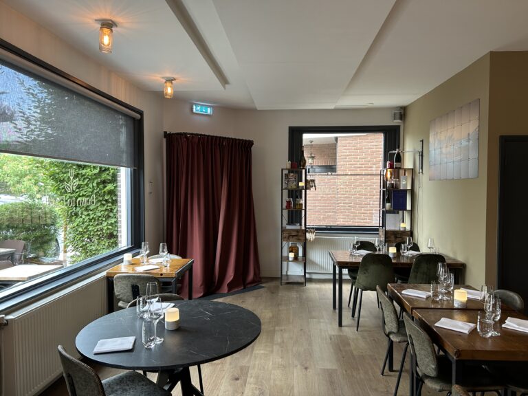 Sfeervol restaurantinterieur met houten en ronde tafels, kaarslicht en een groot raam met de reflectie ‘For Food’.