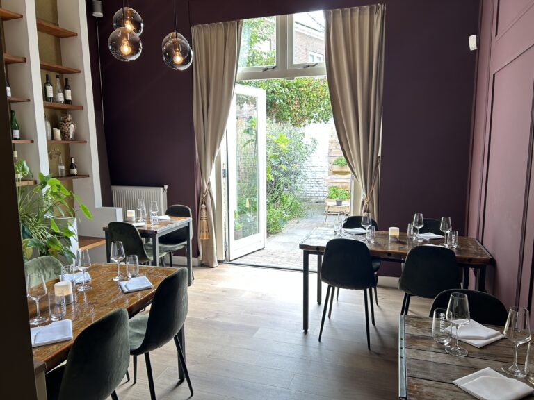 Gezellig restaurantinterieur met donkere muren, houten tafels met groene stoelen, EARTH glazen en openslaande deuren naar tuin.