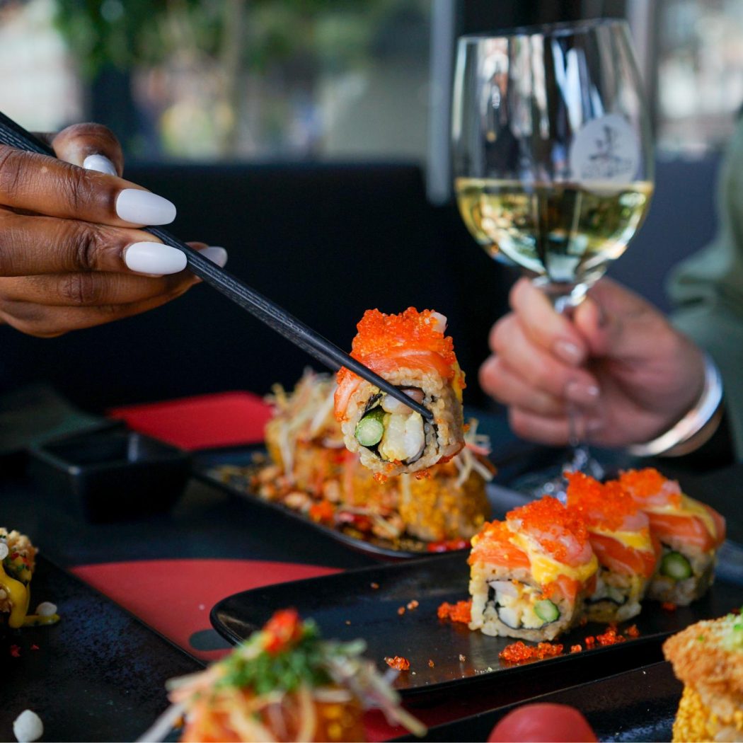 Een hand met witte nagels pakt een sushirol met oranje viseitjes en zalm met zwarte eetstokjes, naast een glas witte wijn.