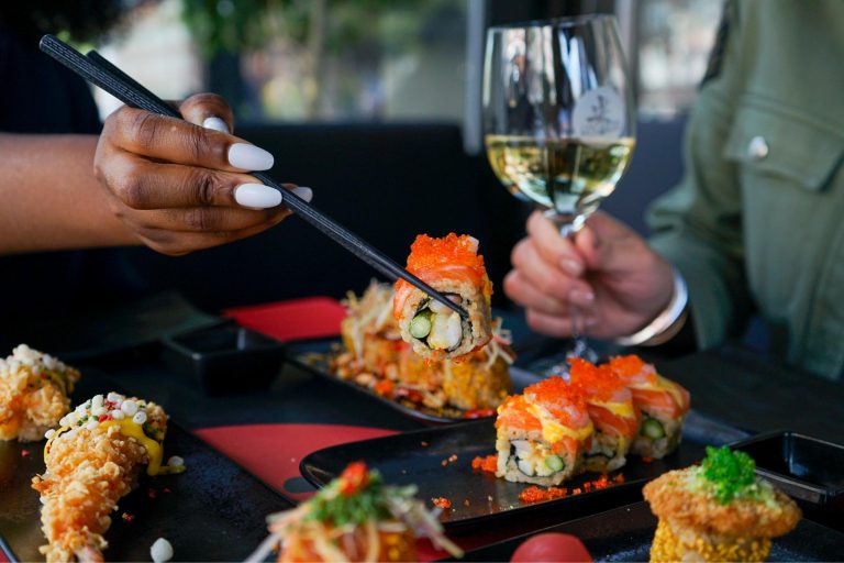 Een hand met witte nagels pakt een sushirol met oranje viseitjes en zalm met zwarte eetstokjes, naast een glas witte wijn.