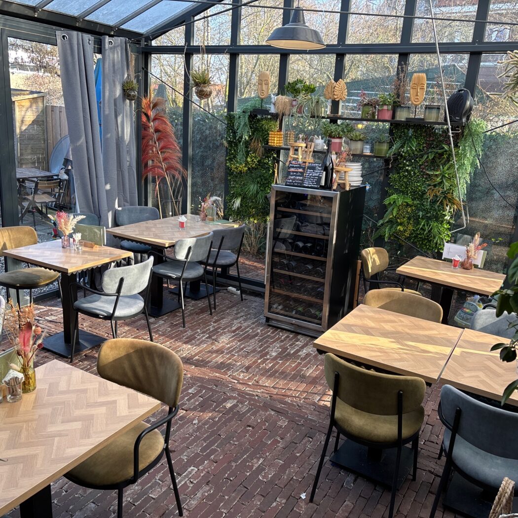 Zonnige serre restaurant met houten tafels, groene en grijze stoelen en een weelderige plantenwand.