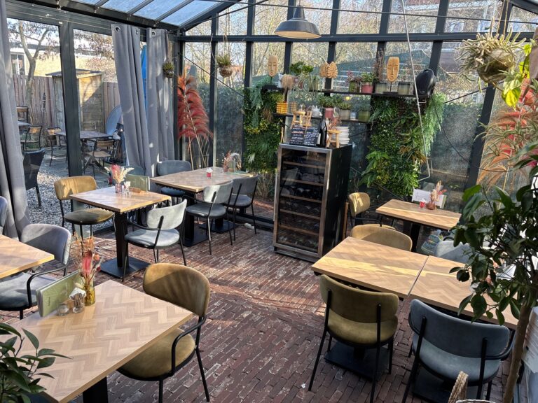 Zonnige serre restaurant met houten tafels, groene en grijze stoelen en een weelderige plantenwand.