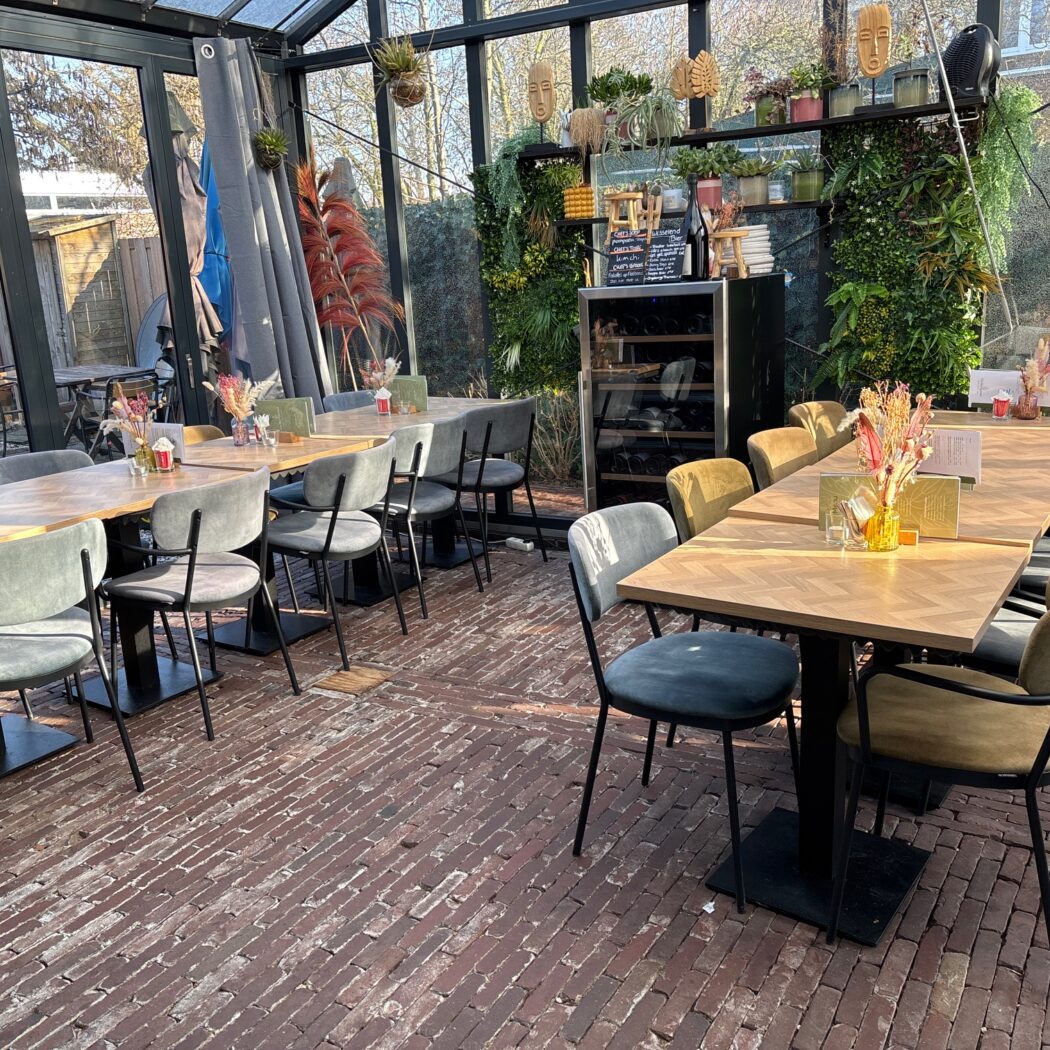 Sfeervolle restaurant serre met houten visgraat tafels, diverse fluwelen stoelen en een groene plantenmuur.