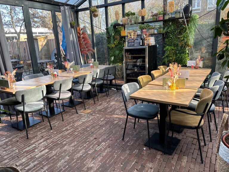 Sfeervolle restaurant serre met houten visgraat tafels, diverse fluwelen stoelen en een groene plantenmuur.