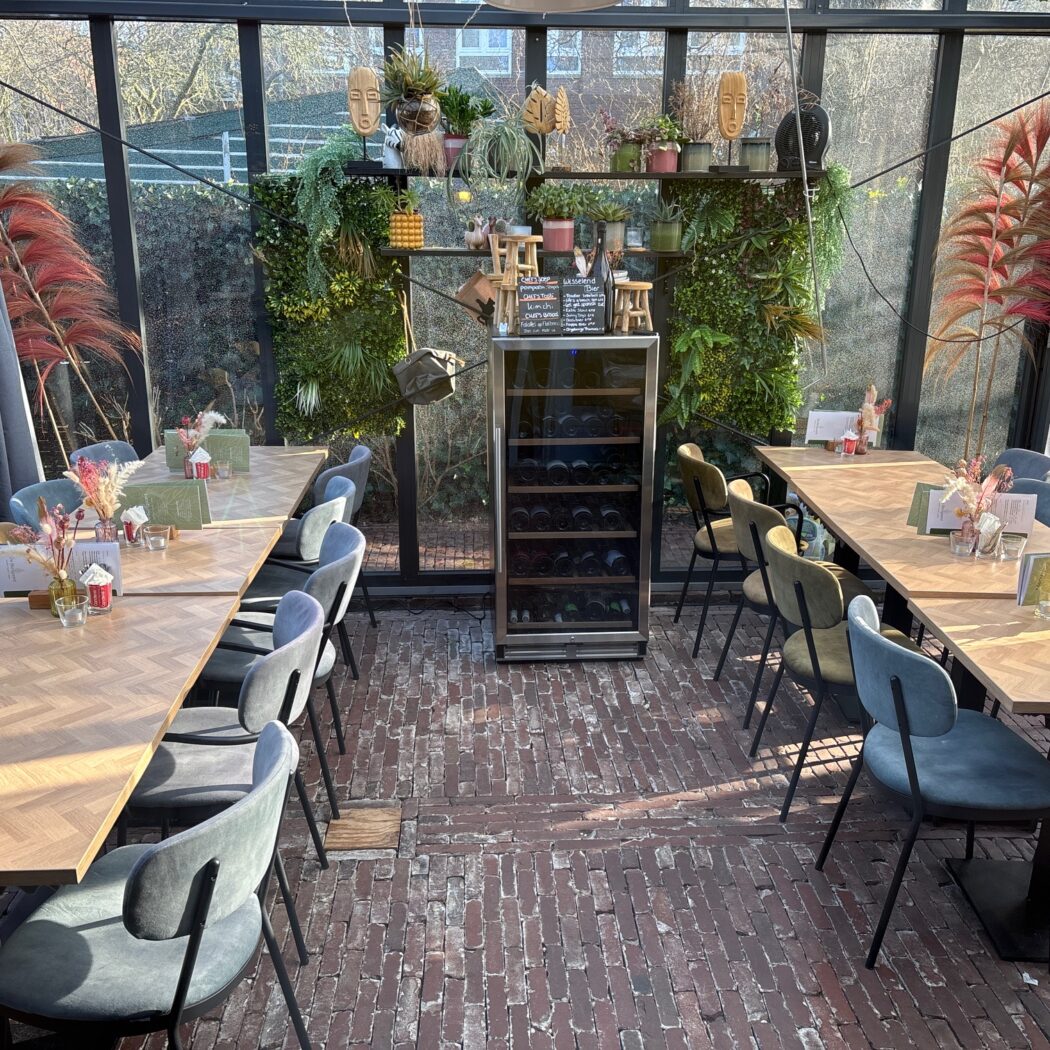 Lichte restaurant serre met houten tafels, gestoffeerde stoelen, een groene plantenwand en unieke decoraties.