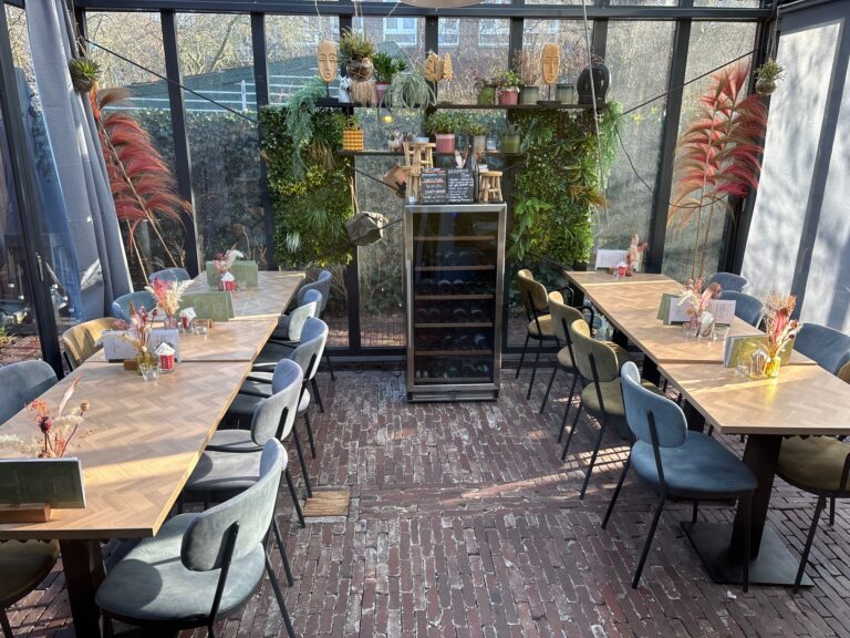 Lichte restaurant serre met houten tafels, gestoffeerde stoelen, een groene plantenwand en unieke decoraties.