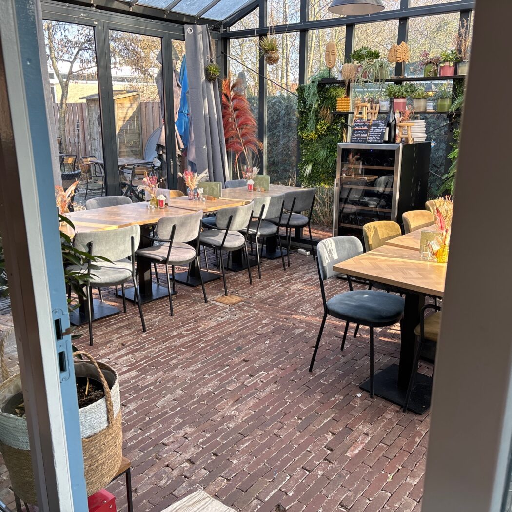 Zonnig restaurant in serre met bakstenen vloer, diverse tafels en een groene plantenwand.