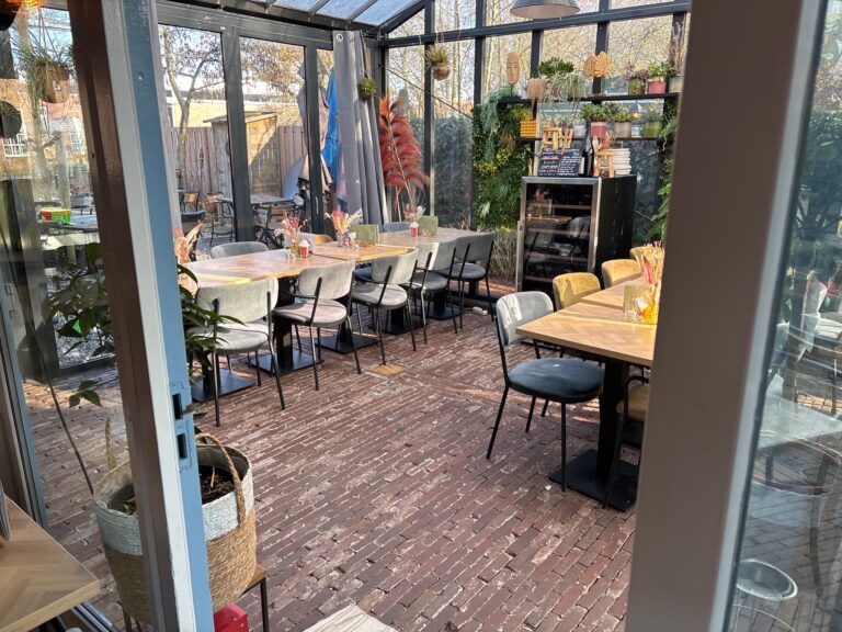 Zonnig restaurant in serre met bakstenen vloer, diverse tafels en een groene plantenwand.