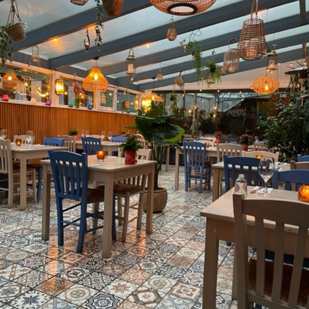 Sfeervol restaurant met houten tafels, blauwe stoelen, kleurrijke mozaïektegels en rieten hanglampen.