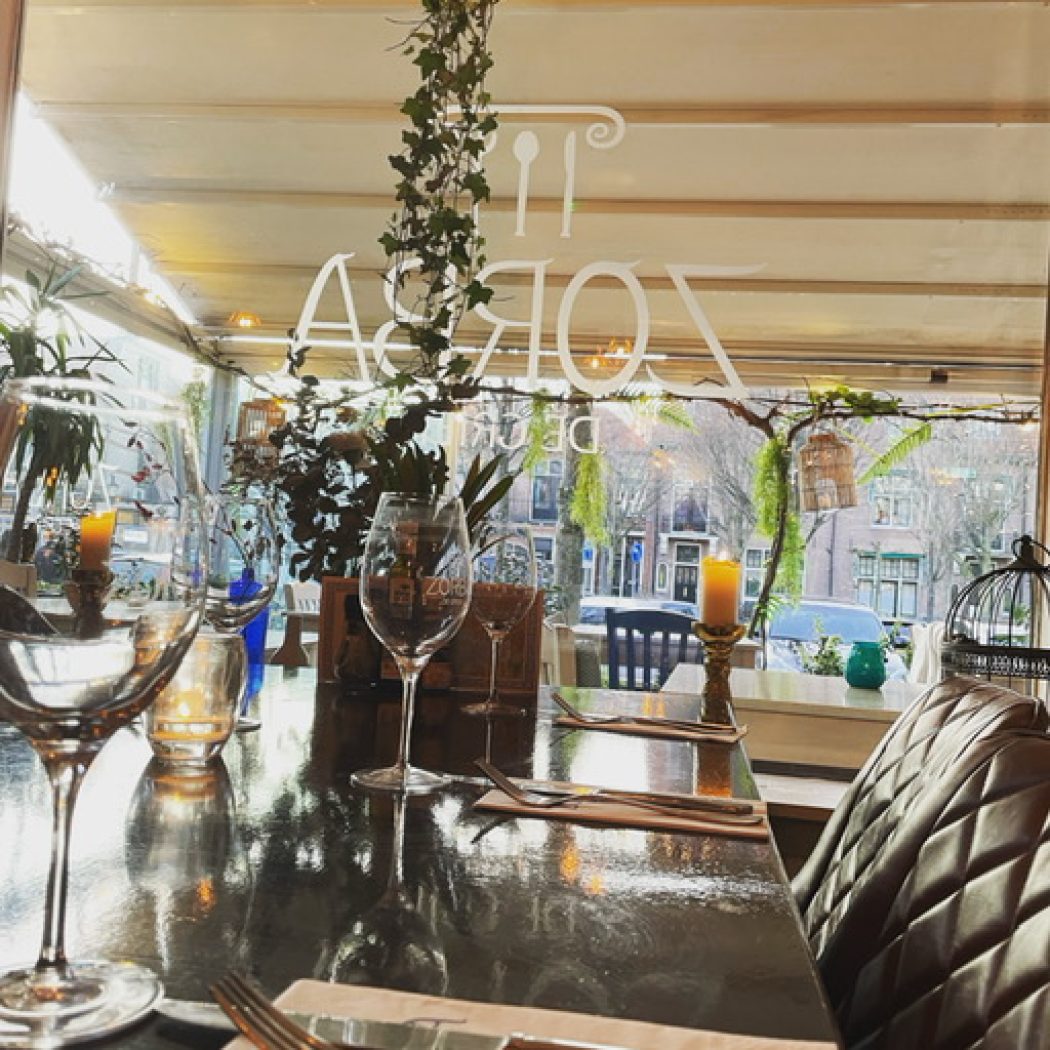 Gedekte restauranttafel met wijnglazen, bestek en brandende kaarsjes; door het raam zie je de straat en omgekeerde tekst IT ZORBA.