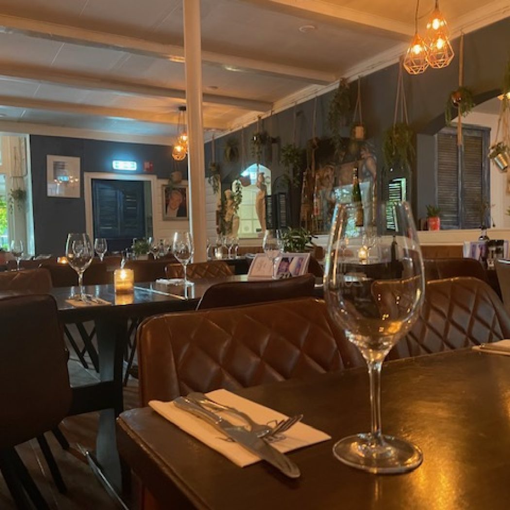 Donker restaurantinterieur met bruine, gewatteerde leren stoelen, gedekte tafels en hangplanten onder sfeervolle verlichting.