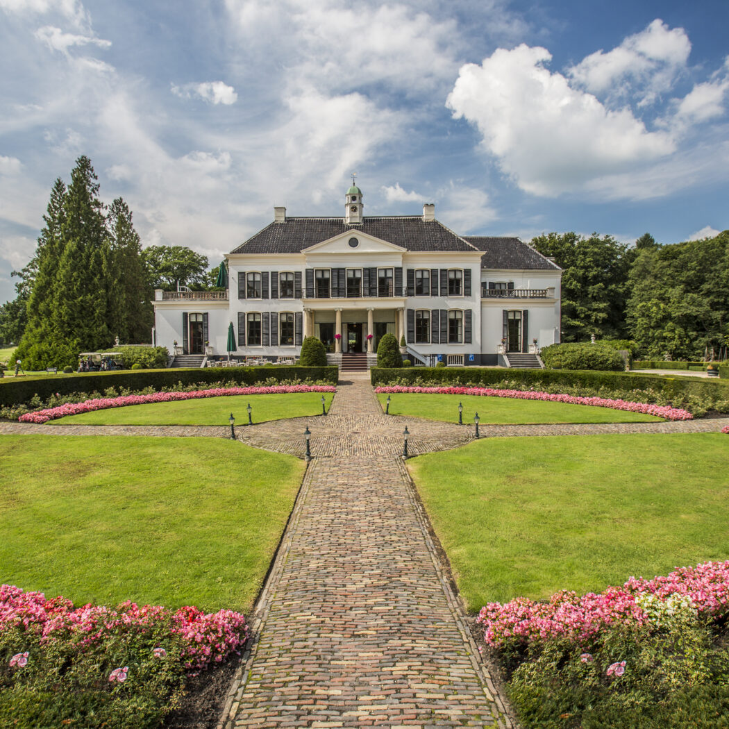 Statig wit landhuis met donkere luiken en een bakstenen pad door een tuin met roze bloembedden.