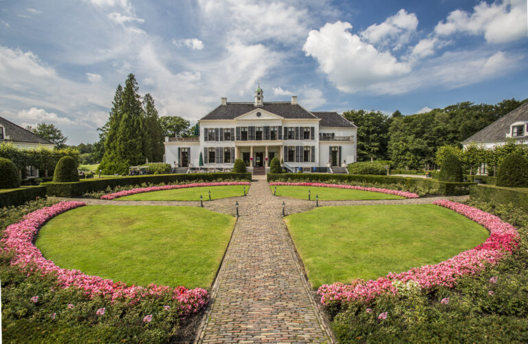 Statig wit landhuis met donkere luiken en een bakstenen pad door een tuin met roze bloembedden.