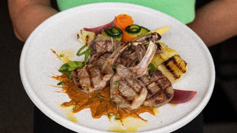 Gegrilde lamskoteletten met gegrilde ananas, courgette, wortel en rode saus op een wit bord.