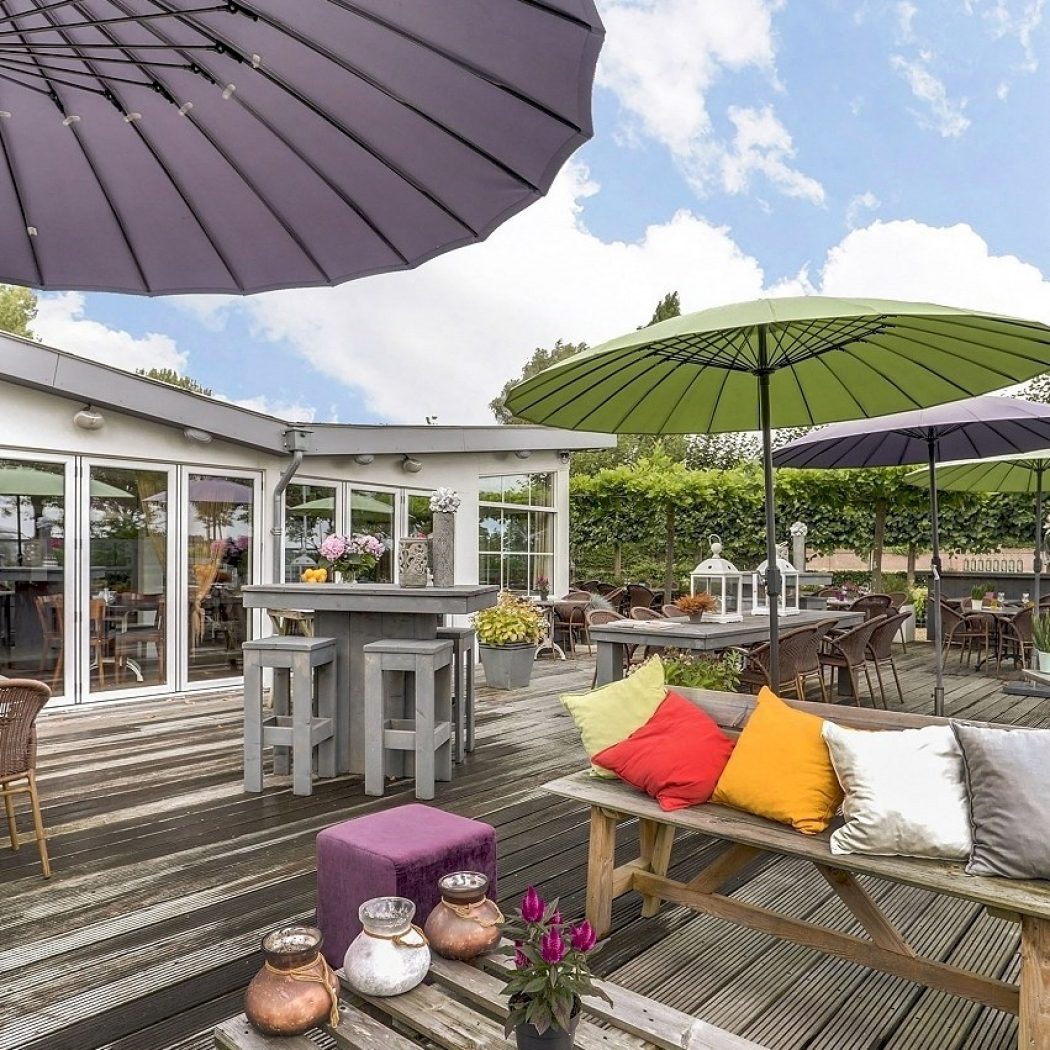 Gezellig buitenterras met houten vlonder, diverse zitjes, kleurrijke kussens en parasols onder een blauwe lucht.