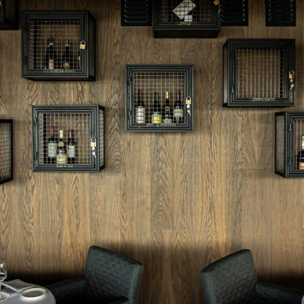 Zwarte metalen wijnkastjes met hangsloten en namen aan een houten wand boven gedekte restauranttafels.