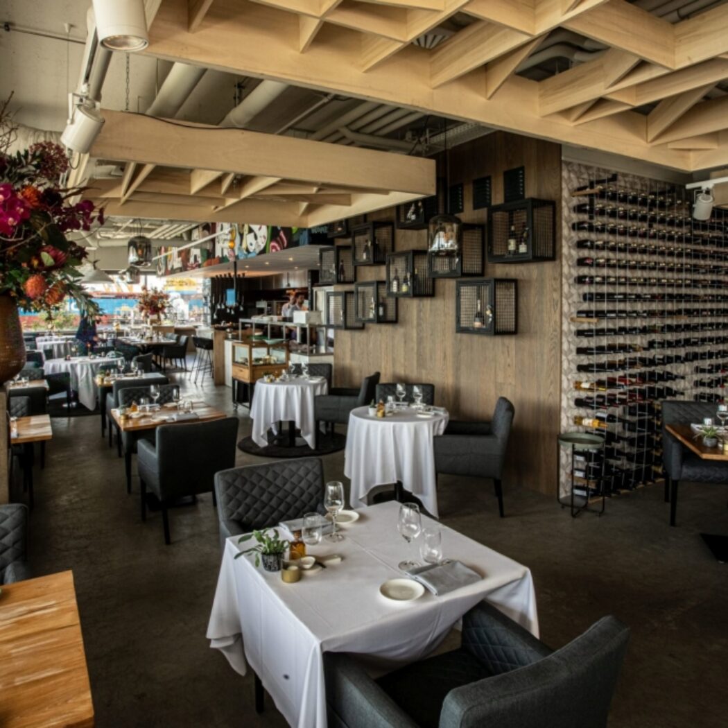 Modern restaurant interieur met gedekte tafels, een groot bloemstuk en een muurvullend wijnrek.