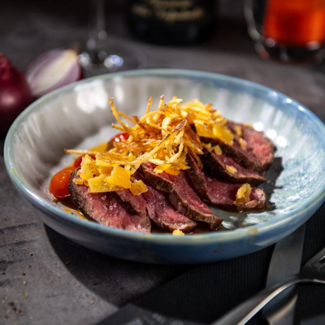 Blauwe kom met dungesneden medium-rare biefstuk, krokante aardappelstros, kaas en cherrytomaten.