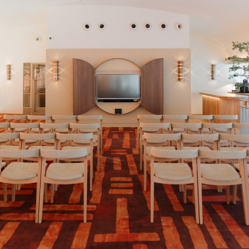 Modern ingerichte evenementenruimte met rijen licht houten stoelen, een rood patroonkleed en scherm omkaderd door houten panelen.