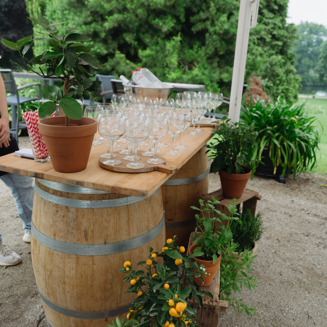 Houten buitenbar van vaten met veel wijnglazen, potplanten met oranje vruchten en een witte parasol in de tuin.