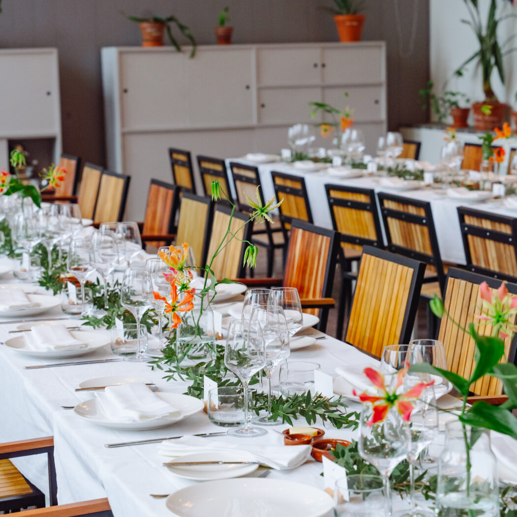 Lange feesttafel gedecoreerd met wit servies, groene bladeren, oranje bloemen en houten stoelen.
