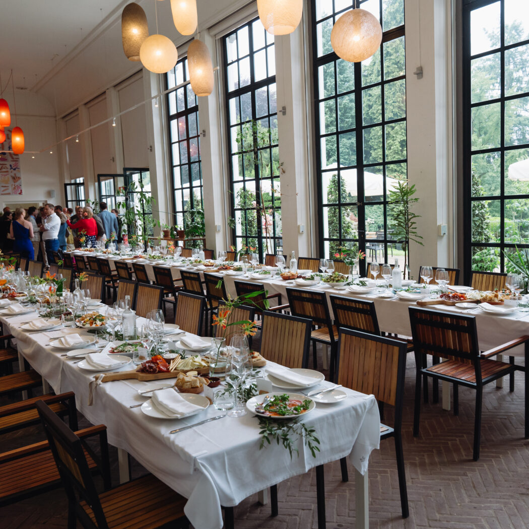 Lange, gedekte tafels voor een evenement in een lichte zaal met hoge ramen en groen uitzicht, met gasten op de achtergrond.