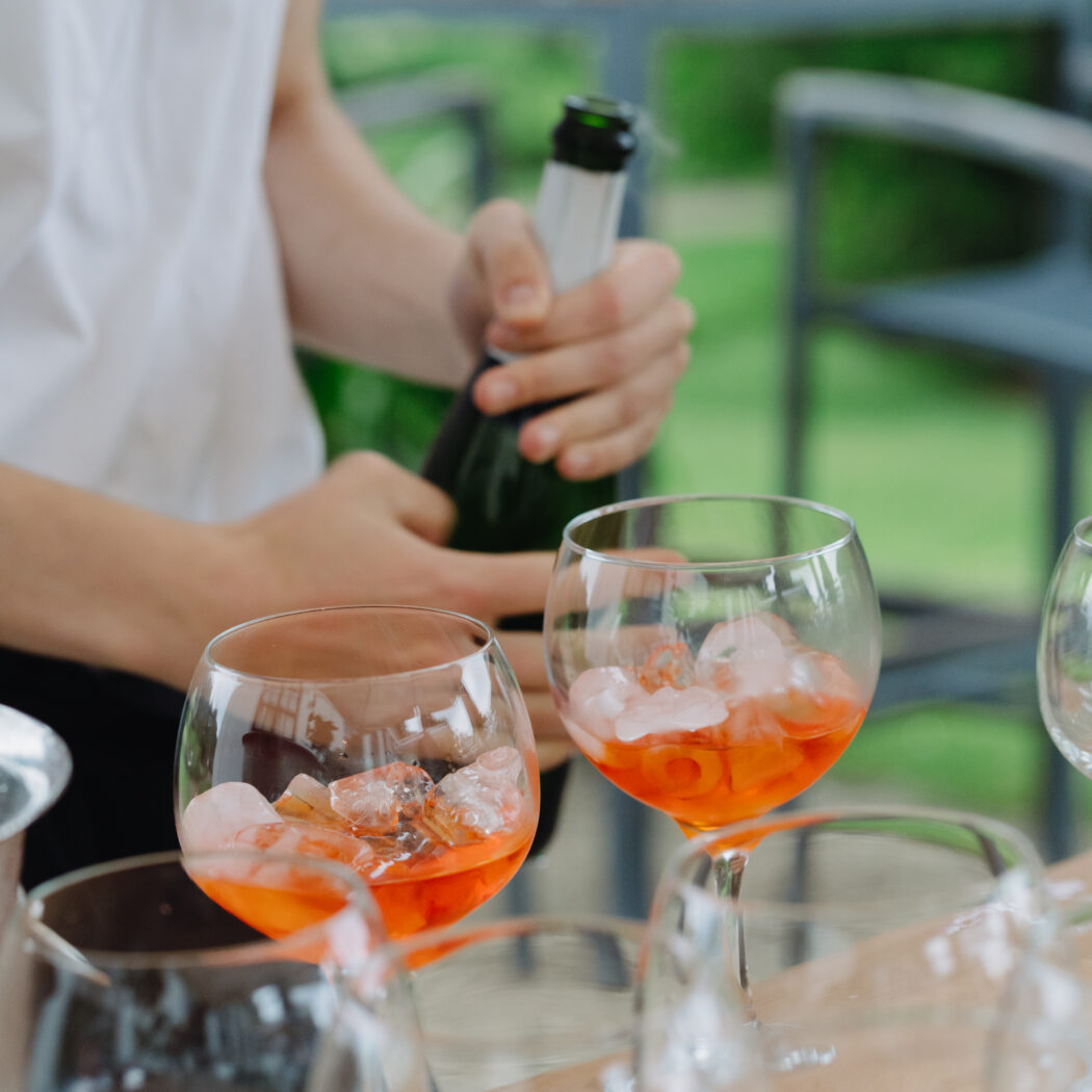 Mousserende wijnfles wordt ontkurkt naast twee oranje cocktails met ijsblokjes op een houten tafel.