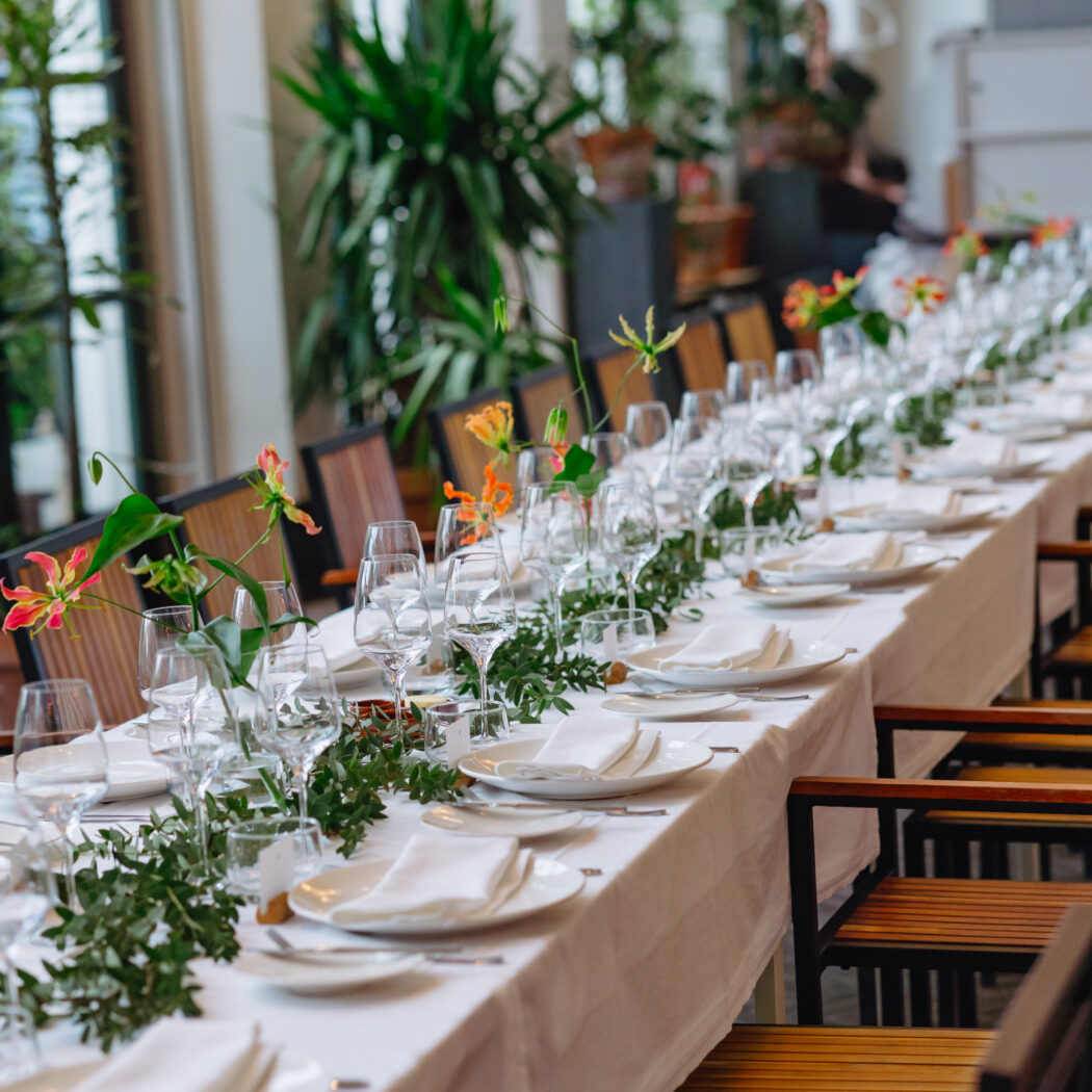 Lange gedekte tafel met witte borden, glazen, groene bladguirlande en oranje bloemen in een lichte zaal.