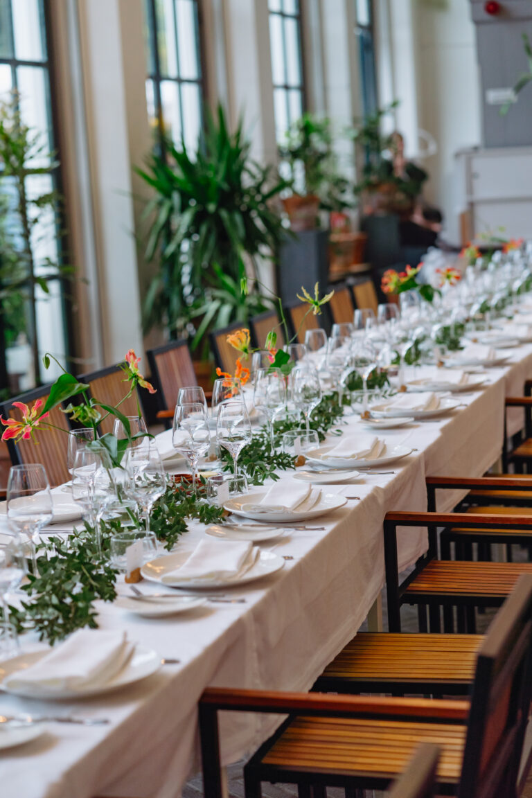 Lange gedekte tafel met witte borden, glazen, groene bladguirlande en oranje bloemen in een lichte zaal.