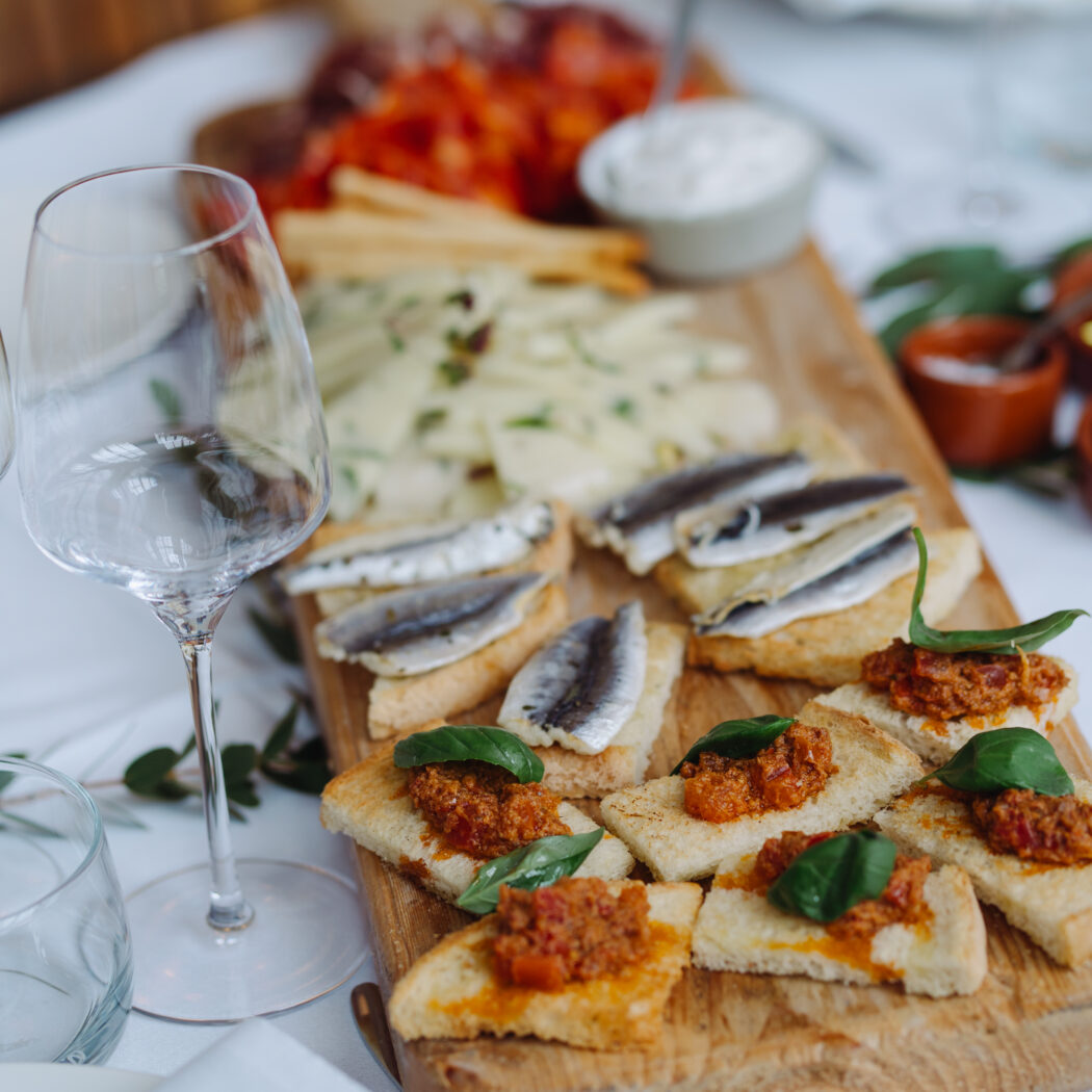 Houten borrelplank met bruschetta met basilicum en toastjes met sardines op een witte tafelloper, naast een wijnglas.