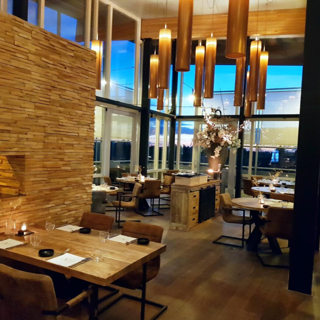Modern restaurantinterieur met houten lambrisering, tafels, stoelen en sfeervolle hanglampen bij avond.