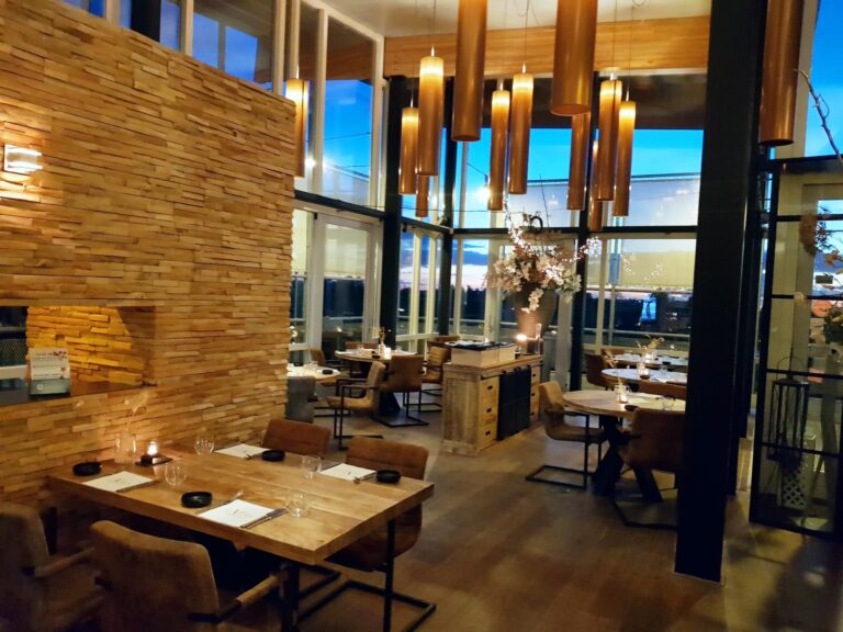 Modern restaurantinterieur met houten lambrisering, tafels, stoelen en sfeervolle hanglampen bij avond.