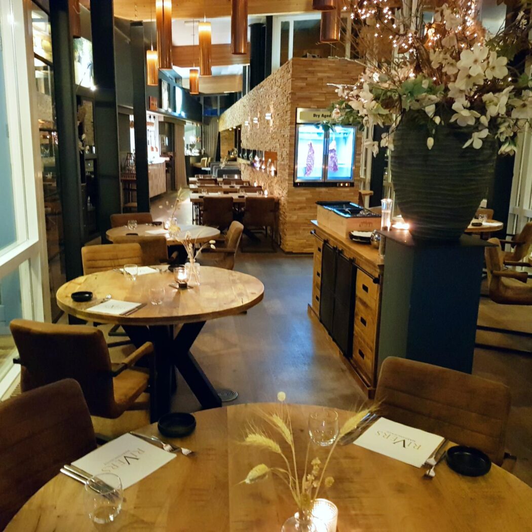 Sfeervol restaurant met ronde houten tafels, bruine stoelen en warme sfeerverlichting. Grote bloemenvaas, Dry Aged display en RIVERS menus.