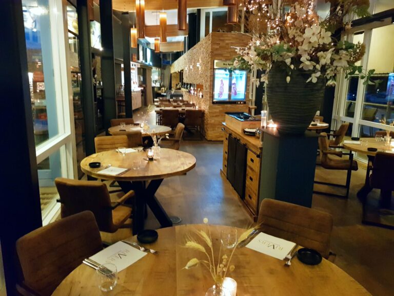 Sfeervol restaurant met ronde houten tafels, bruine stoelen en warme sfeerverlichting. Grote bloemenvaas, Dry Aged display en RIVERS menus.