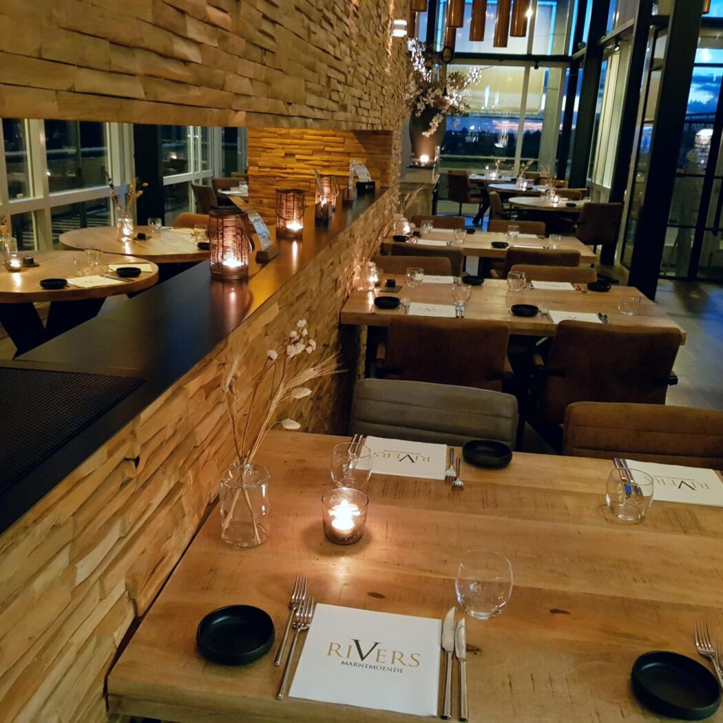 Sfeervol restaurantinterieur met gedekte houten tafels, brandende kaarsen, open haard en RIVERS placemats.