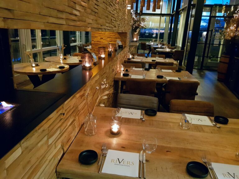 Sfeervol restaurantinterieur met gedekte houten tafels, brandende kaarsen, open haard en RIVERS placemats.