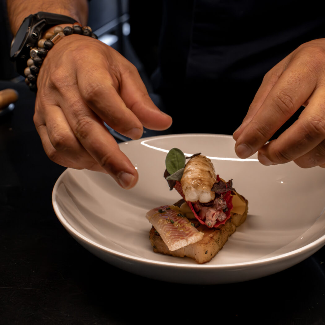 Chef met horloge en kralenarmbanden dresseert een visgerecht met zeevruchten op een witte schaal.