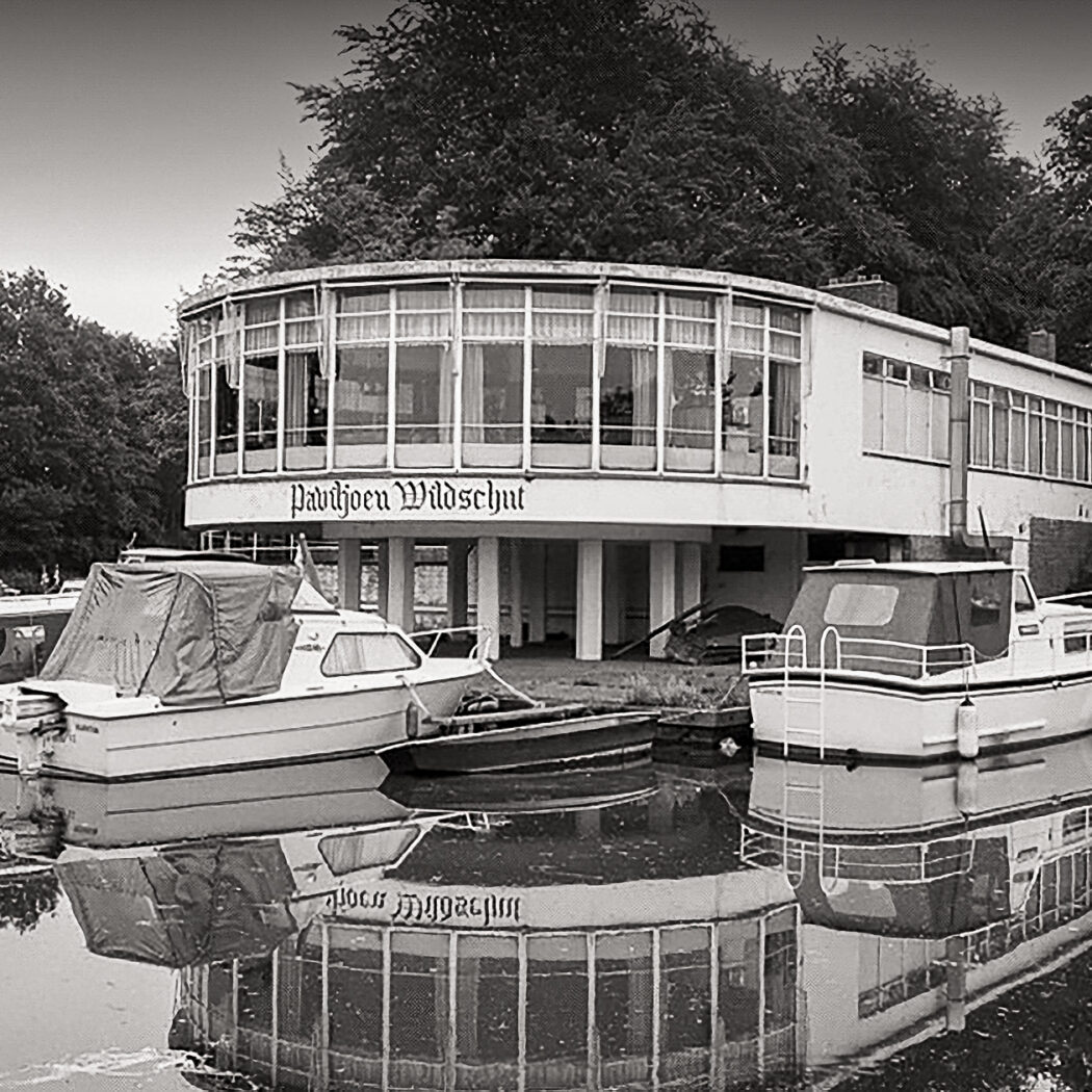 Zwart-witbeeld van Paviljoen Wildschut, een gebouw met veel ramen, met boten en weerspiegelingen in het water.