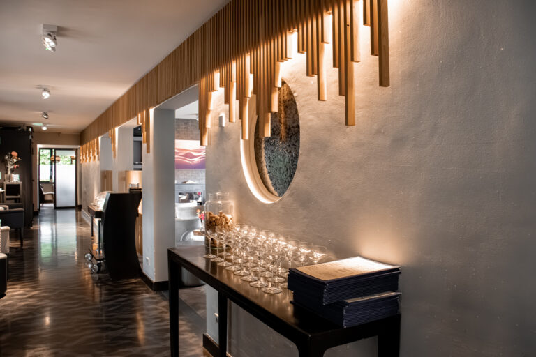 Elegante hal met houten lamellen plafondverlichting, ronde muuruitsparing en een tafel met rijen glazen en menukaarten.