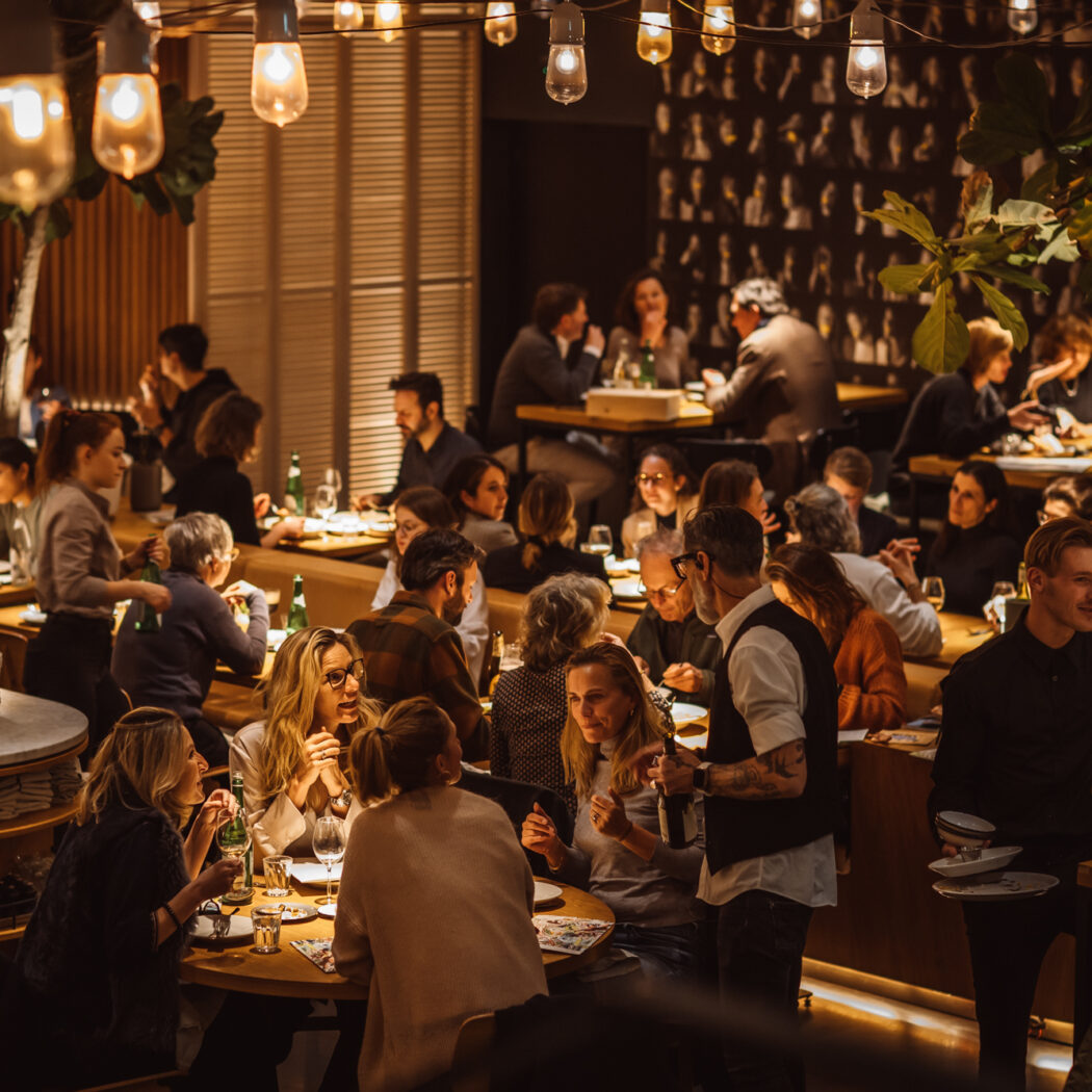 Drukke horecazaak met veel gasten aan tafel, bedienend personeel en warme sfeerverlichting.